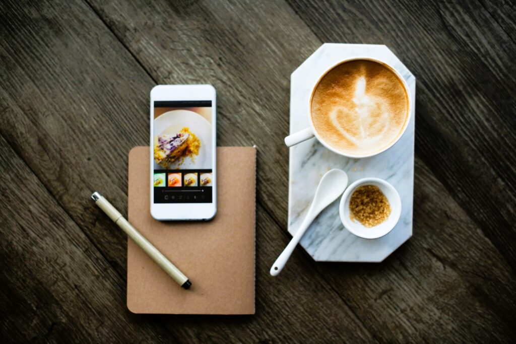 A smartphone displaying a food photo editing app rests on a rustic wooden table beside a notebook and pen, with a latte and sugar on a marble coaster nearby. This image illustrates how technology can enhance and streamline everyday tasks, even in relaxing settings like enjoying a coffee