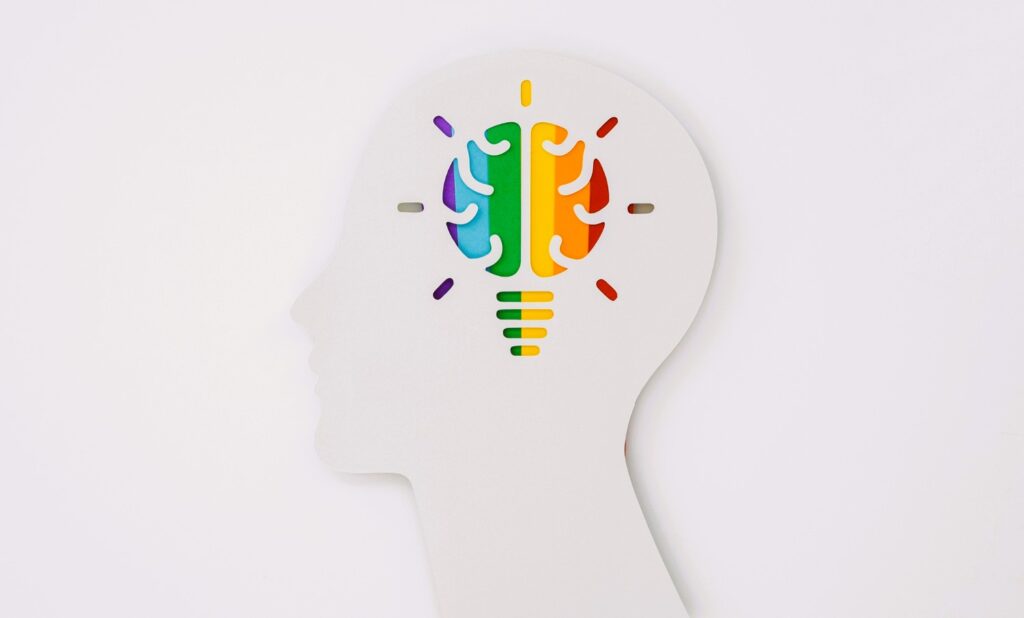 A white silhouette of a human head in profile with a colorful, rainbow-striped brain and lightbulb cut out, symbolizing the connection between creativity, innovation, and cognitive function