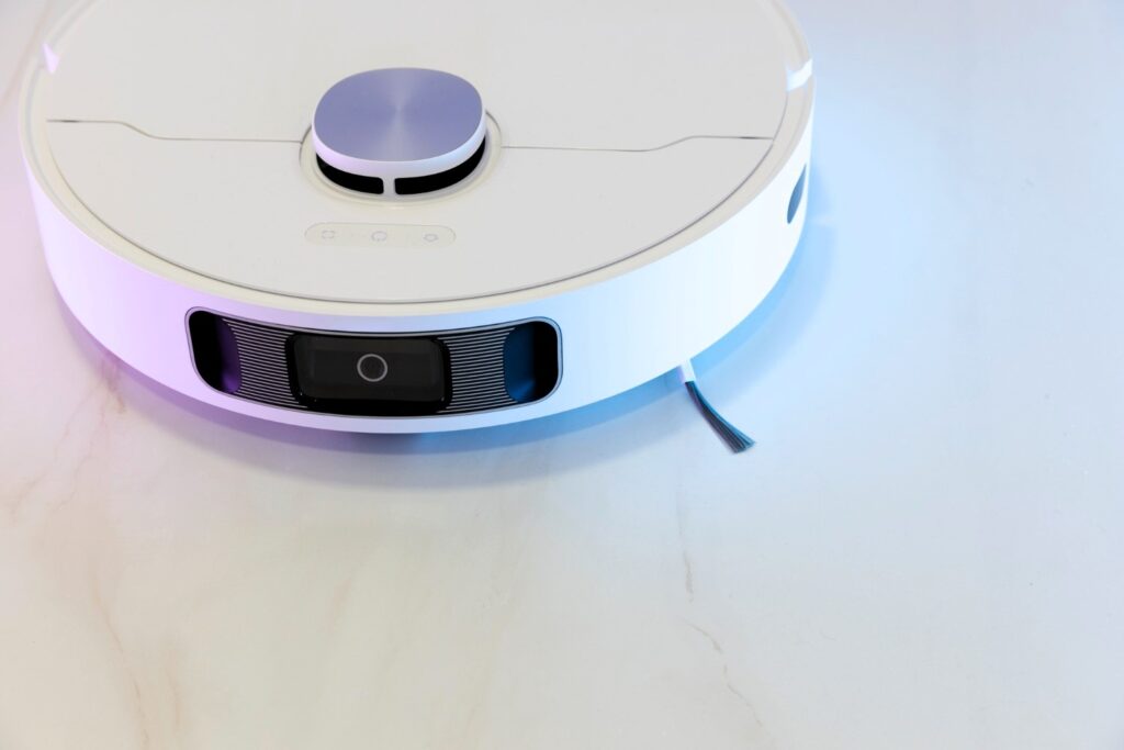 A white robot vacuum cleaner with a sleek design, featuring a top-mounted laser sensor and a front camera for navigation, sits on a marble floor with a stray string tangled in its brush