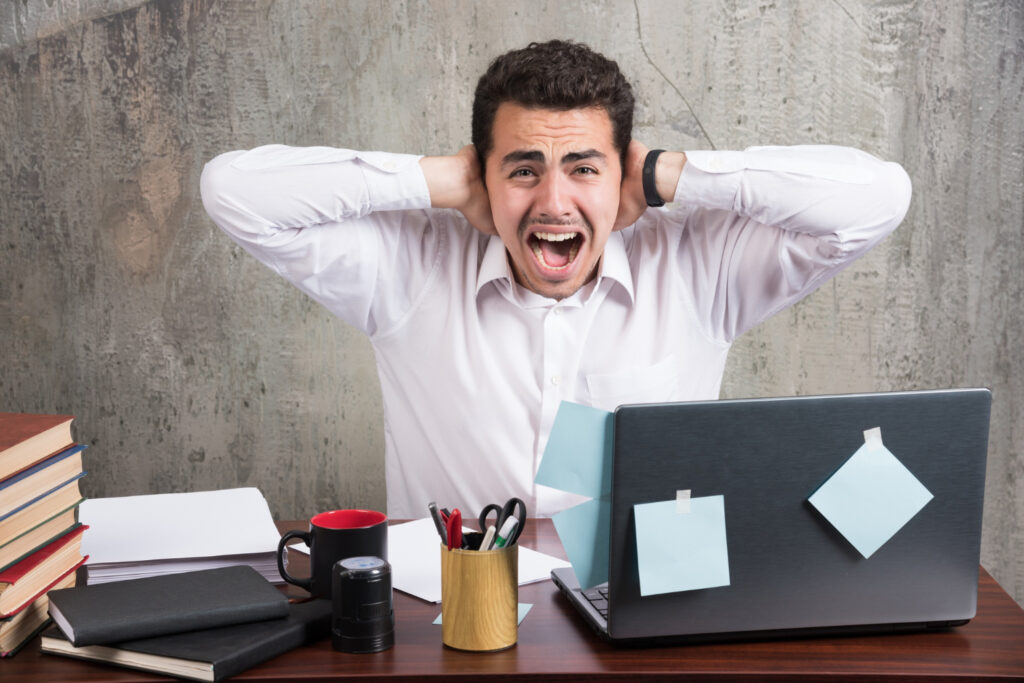 Home Tech Productivity - Office employee screaming while holding his ears at the office desk.