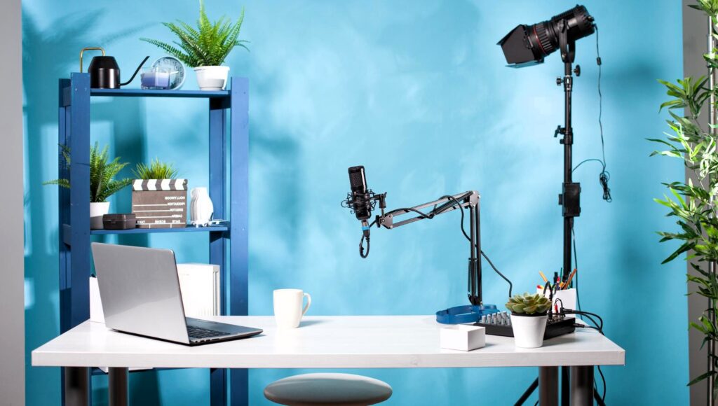 A home podcast or video recording setup with a white desk against a blue wall. The desk holds a laptop, microphone on an adjustable arm, coffee mug, and plant. A shelf behind the desk displays plants, a director's clapboard, and other items. A camera on a tripod and a lighting fixture are set up to record the person sitting at the desk