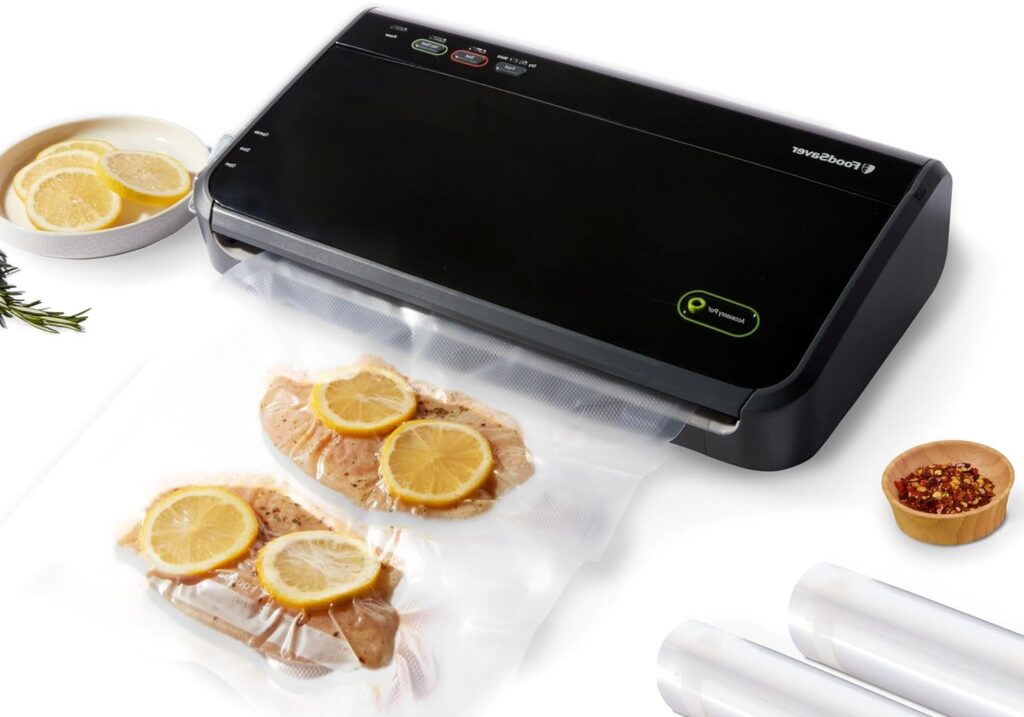 A black FoodSaver vacuum sealer machine on a countertop, sealing two bags of chicken breasts with lemon slices. Rolls of vacuum sealer bags and a bowl of spices are also visible