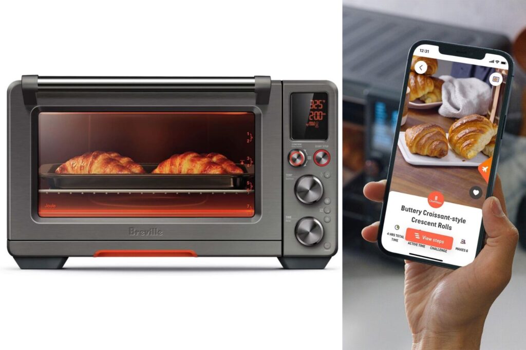 A stainless steel countertop oven with a black door sits on a white counter. A chrome handle is on the door. A smartphone with a recipe on the screen rests on the counter beside the oven. Text on the Breville countertop oven reads "Breville Smart Oven Air Fryer Pro with Connected App